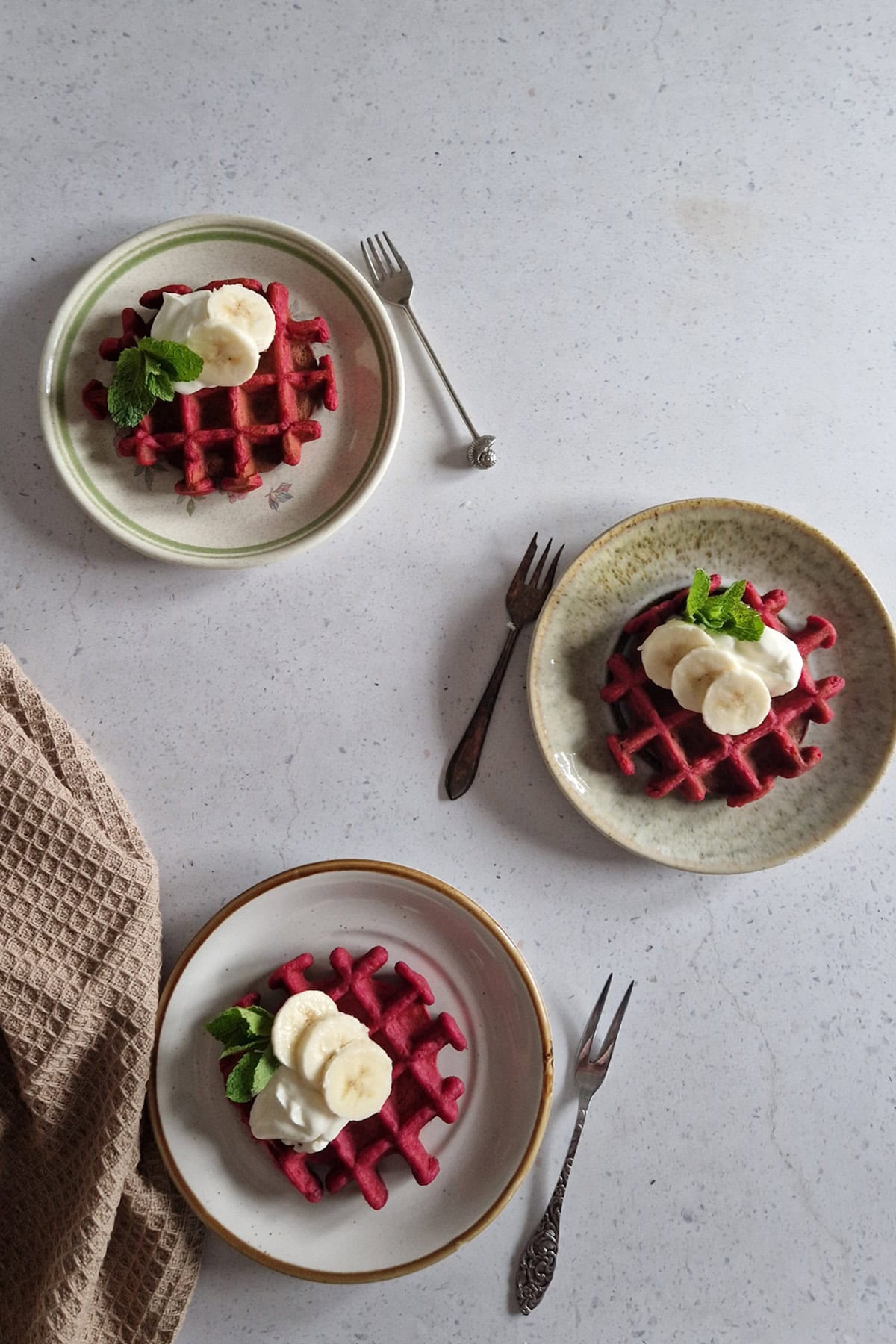 3 beet waffles on small plates, with yoghurt and banana as a topping.