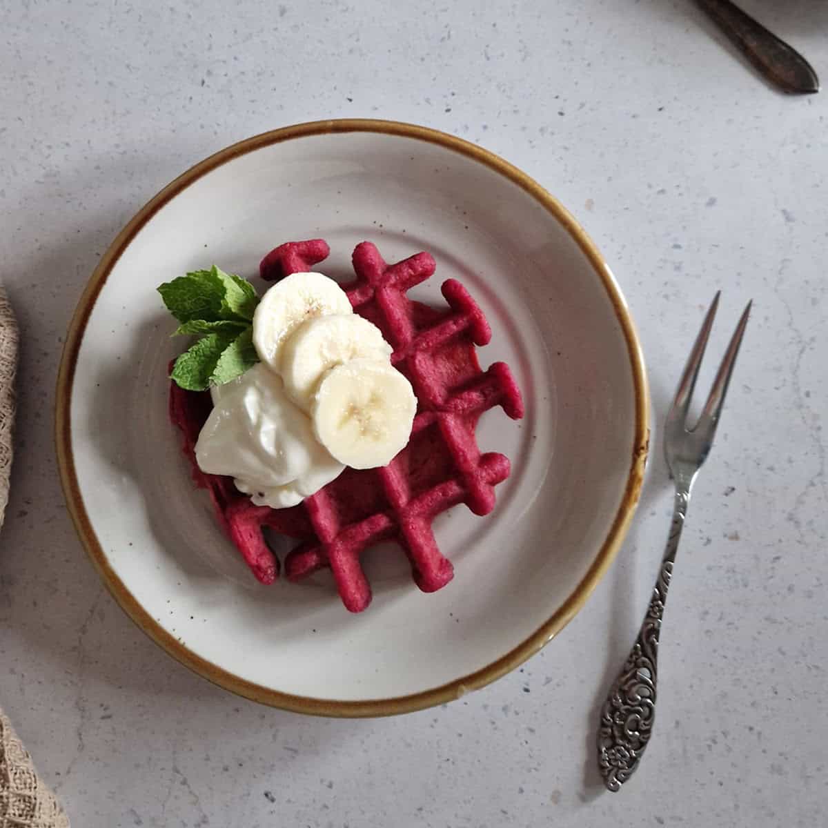 A bright pink waffle on a little plate, topped with a dollop of yoghurt and some slices of banana.