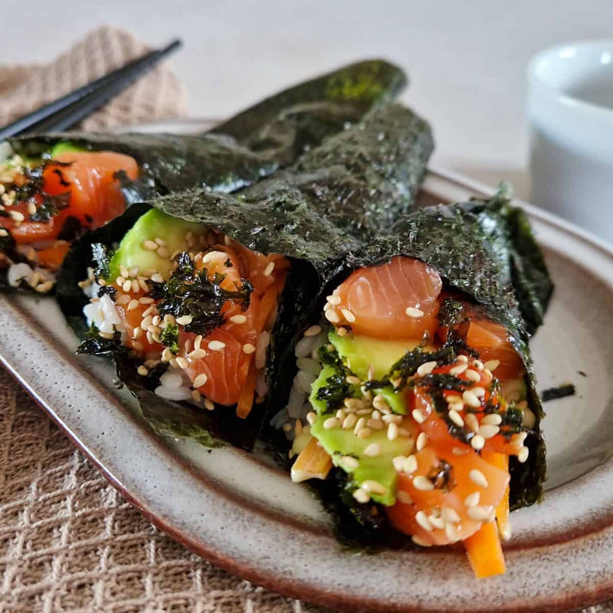 Temaki sushi filled with salmon and avocado, on a plate. The hand rolls are garnished with sesame seeds.