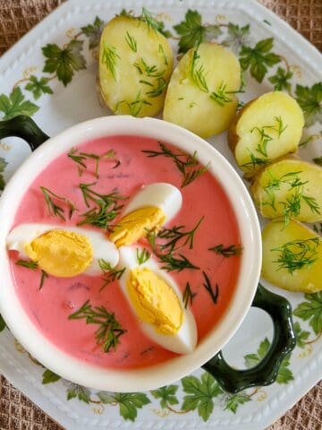 A bowl of Šaltibarščiai (Lithuanian Cold Beet Soup), with a side of cooked potatoes and dill. The dish is served on an old-fashioned green and white plate.