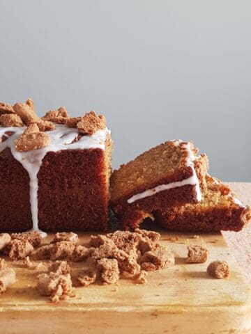 A cinnamon loaf cake with a few slices cut off. The cinnamon cake is covered with a white frosting and a cookie crumble topping.