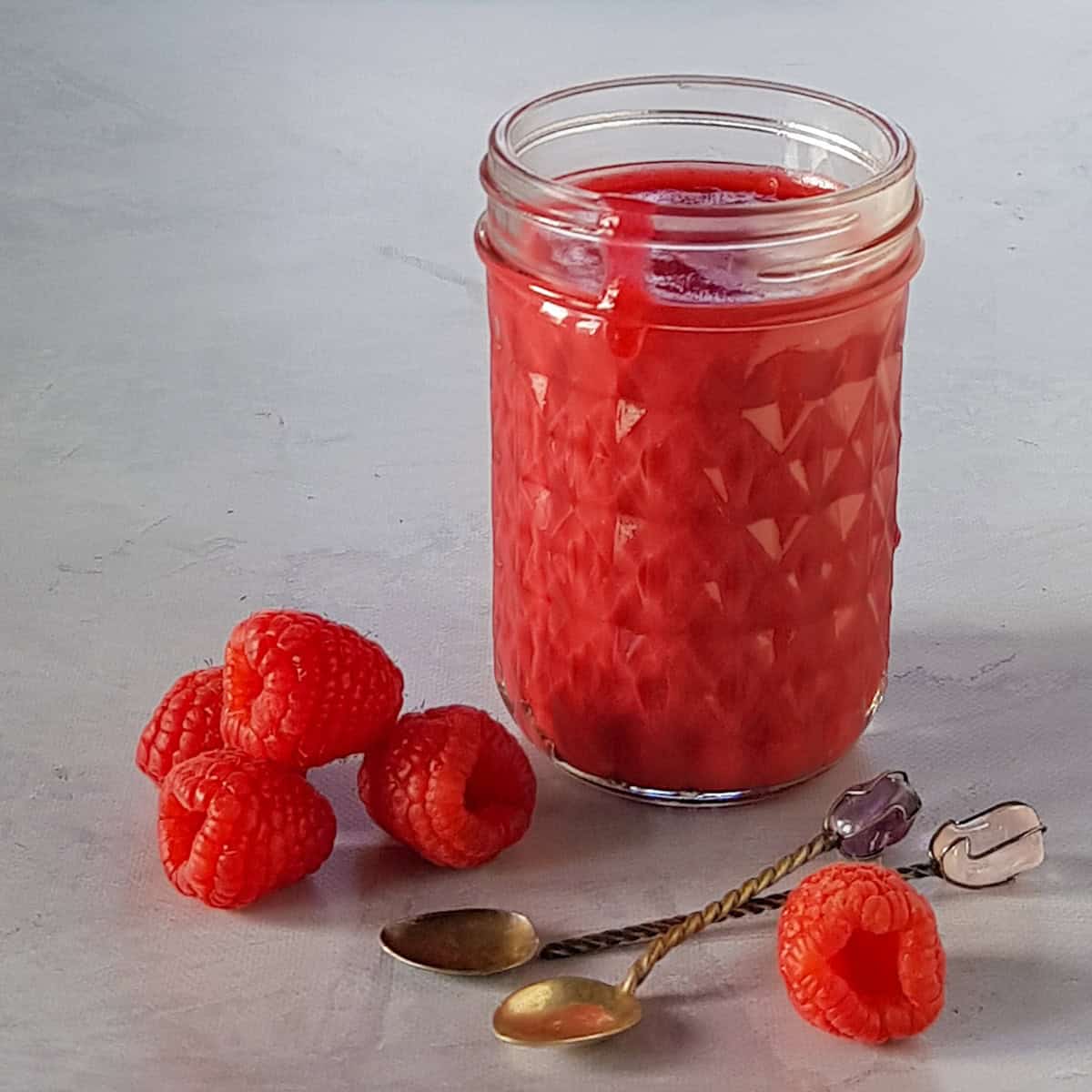 A glass jar filled with homemade raspberry curd, surrounded by fresh raspberries.