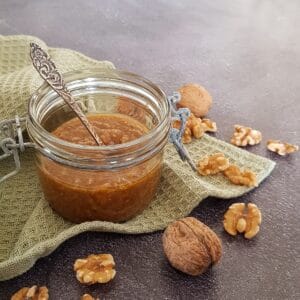 A glass jar filled with homemade walnut pesto. The jar is surrounded by halved and whole walnuts.