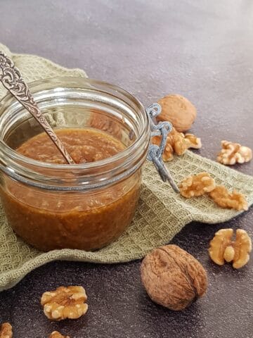 A glass jar filled with homemade walnut pesto. The jar is surrounded by halved and whole walnuts.