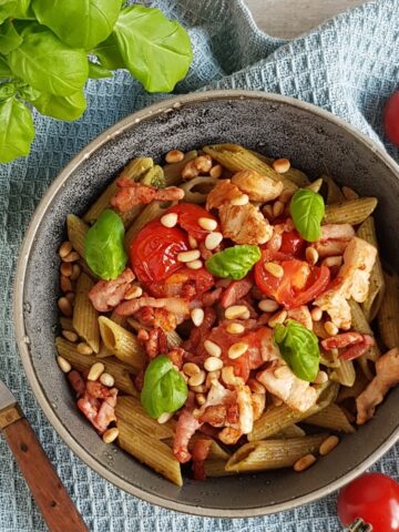 A grey bowl, filled with bacon pesto pasta, topped with tomatoes and fresh basil leaves.