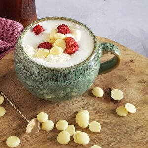 A mug of white chocolate milk, served with white chocolate drops and freeze dried strawberries.