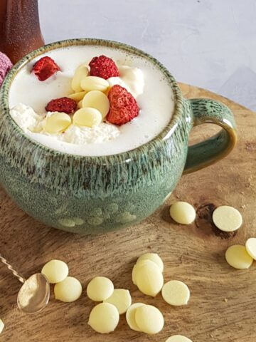 A mug of white chocolate milk, served with white chocolate drops and freeze dried strawberries.