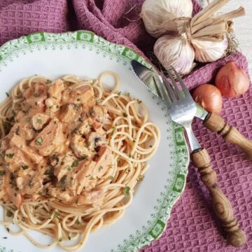 A plate filled with creamy kielbasa pasta, garnished with dried parsley. The plate is surrounded by a fork and knife, garlic bulbs and shallots.