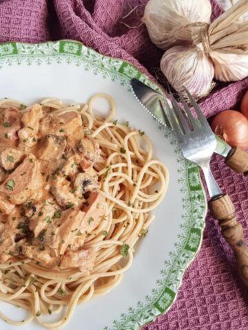 A plate filled with creamy kielbasa pasta, garnished with dried parsley. The plate is surrounded by a fork and knife, garlic bulbs and shallots.