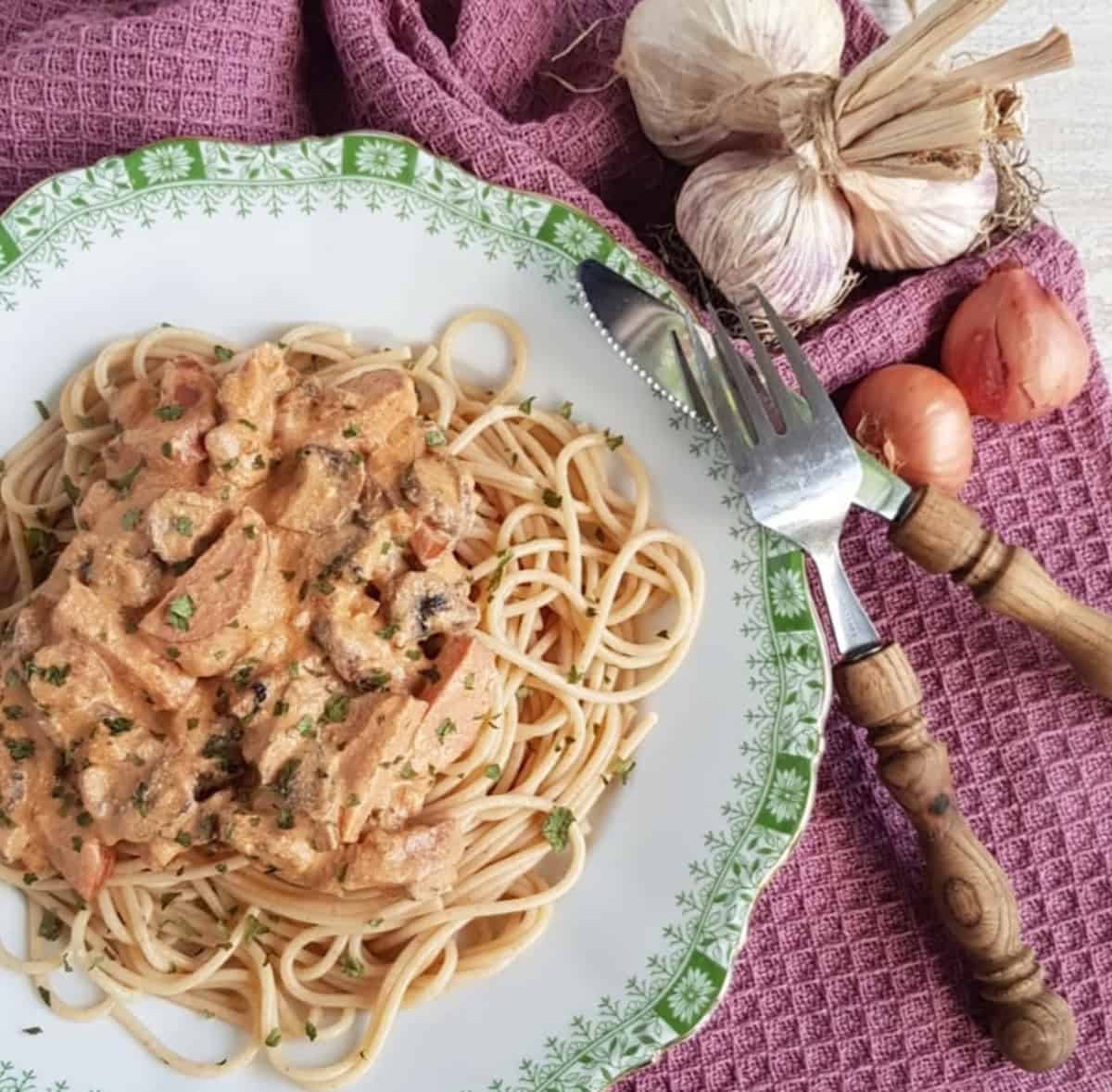 A plate filled with creamy kielbasa pasta, garnished with dried parsley. The plate is surrounded by a fork and knife, garlic bulbs and shallots.