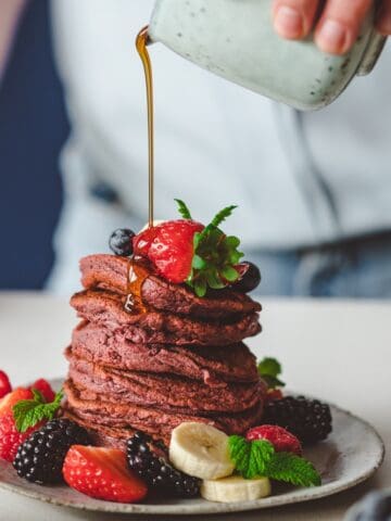A pile of beetroot pancakes covered with fresh fruit. A hand pours syrup over the pink pancakes.