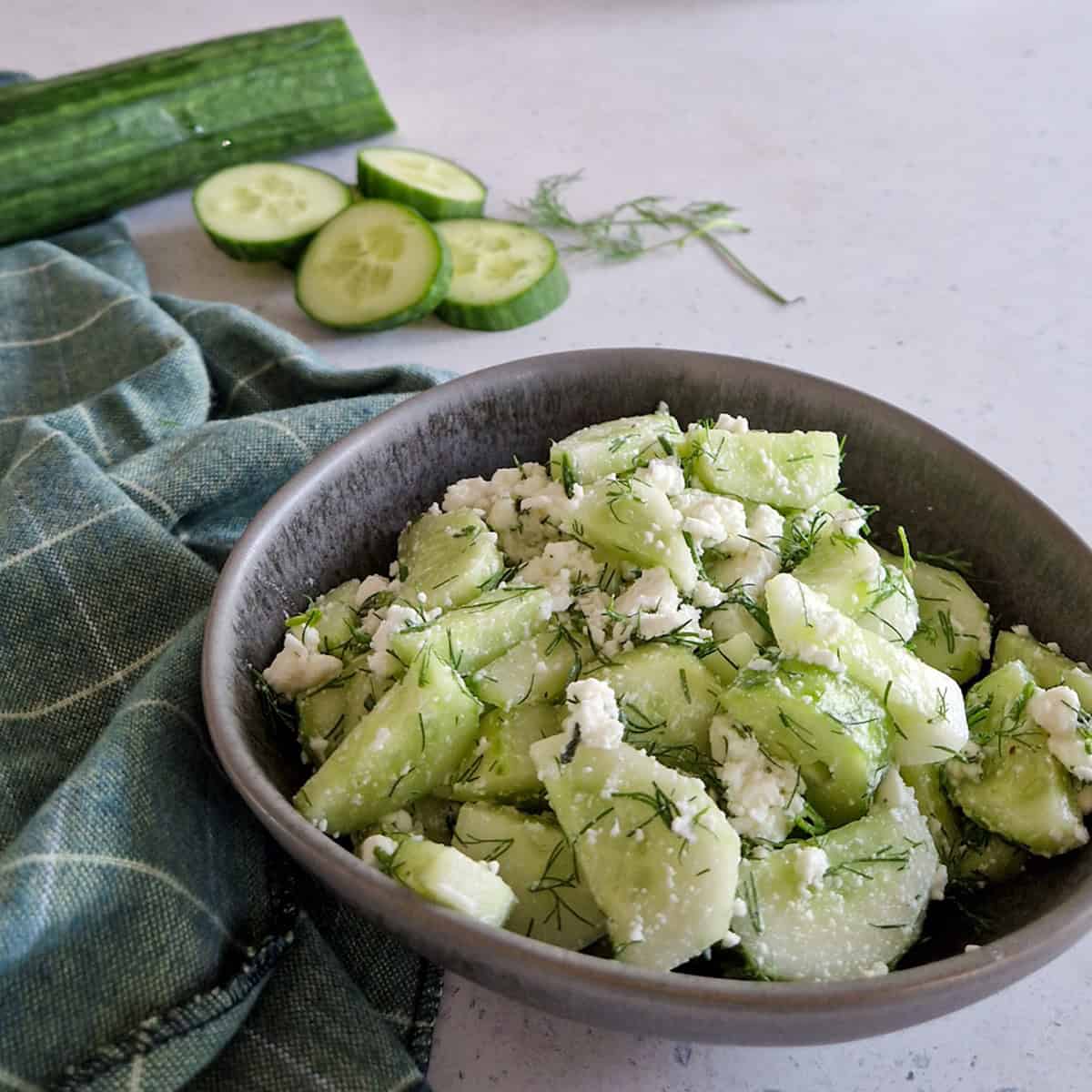 A bowl with a fresh cucumber and dill salad. There are some ingredients of the recipe (cucumber slices and fresh dill) visible in the background.