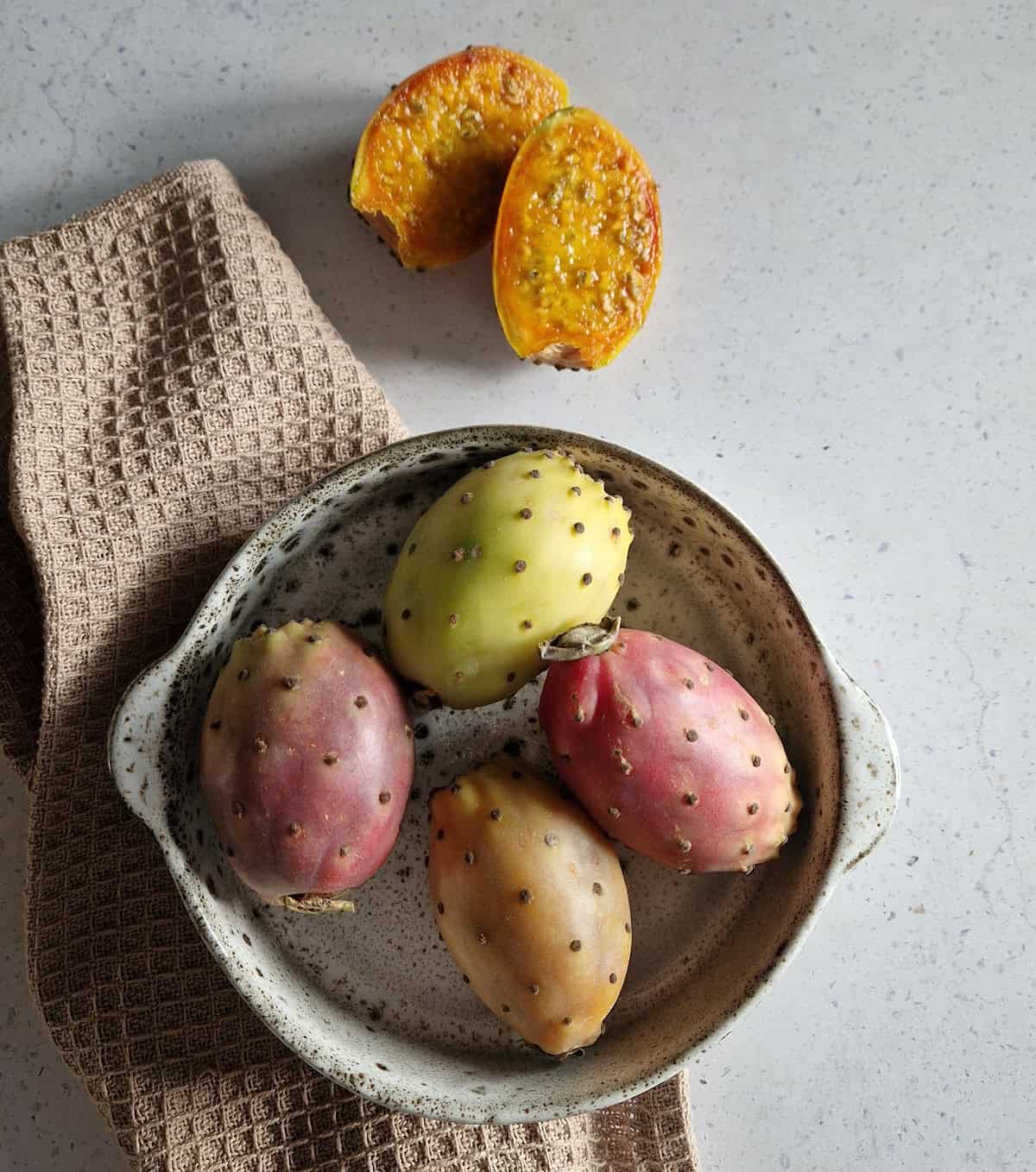 A bowl with four prickly pears (or cactus fruit) in varied colors. One cactus fruit is halved to show the bright orange-yellow inside.