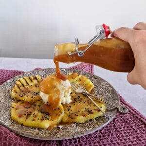 A hand pours soy caramel sauce over a dessert with grilled pineapple and ice cream.