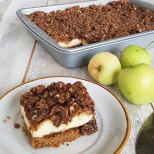 A square of apple crumble cheesecake on a dessertplate. The rest of the cheesecake is still in the baking tin, visible in the background.