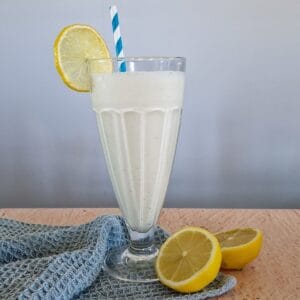 A lemon milkshake in a typical milkshake glass. A halved lemon is lying in front of the glass.