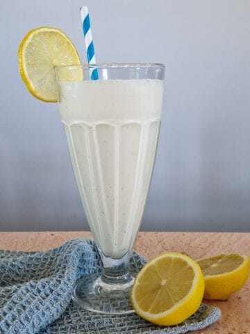 A lemon milkshake in a typical milkshake glass. A halved lemon is lying in front of the glass.