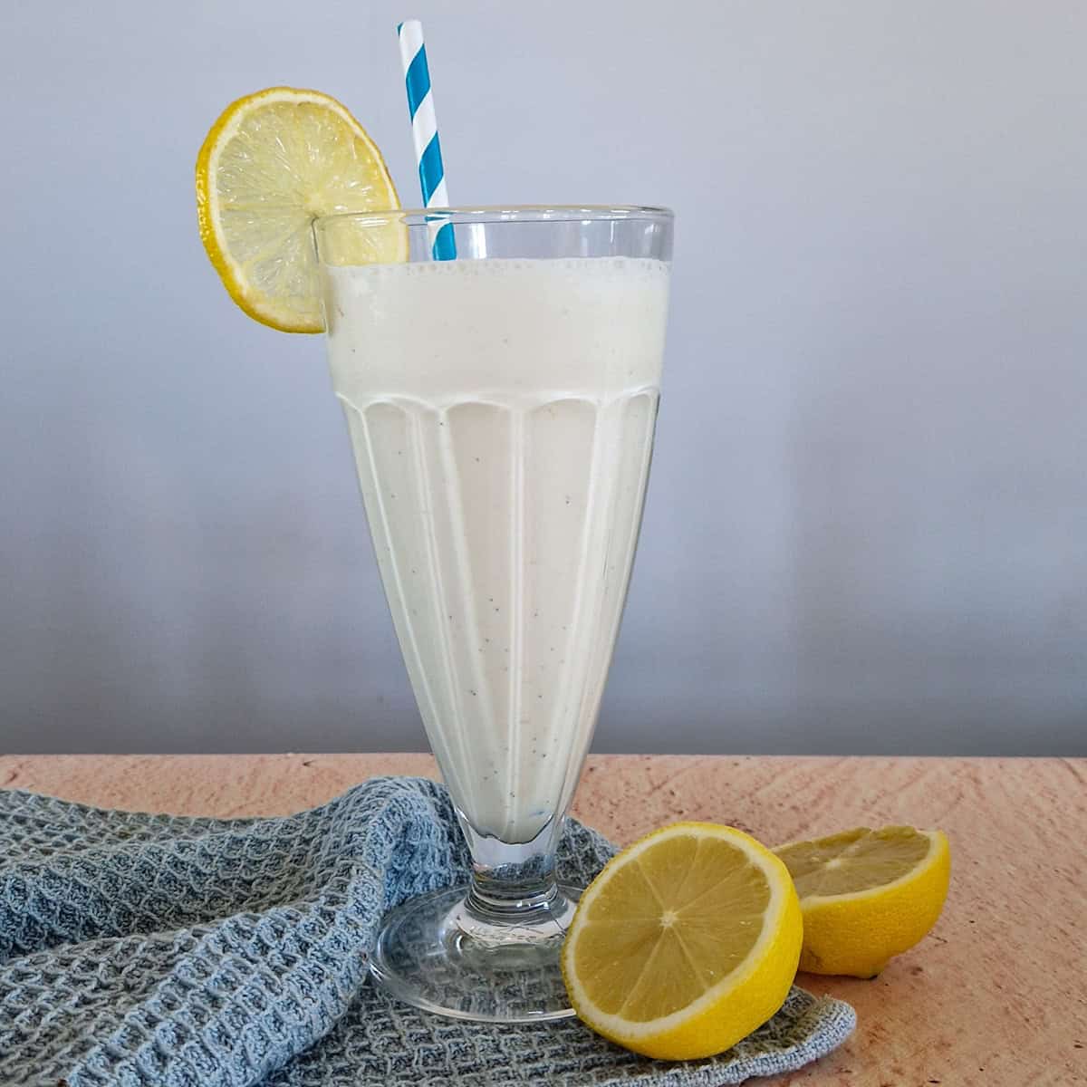 A lemon milkshake in a typical milkshake glass. A halved lemon is lying in front of the glass.