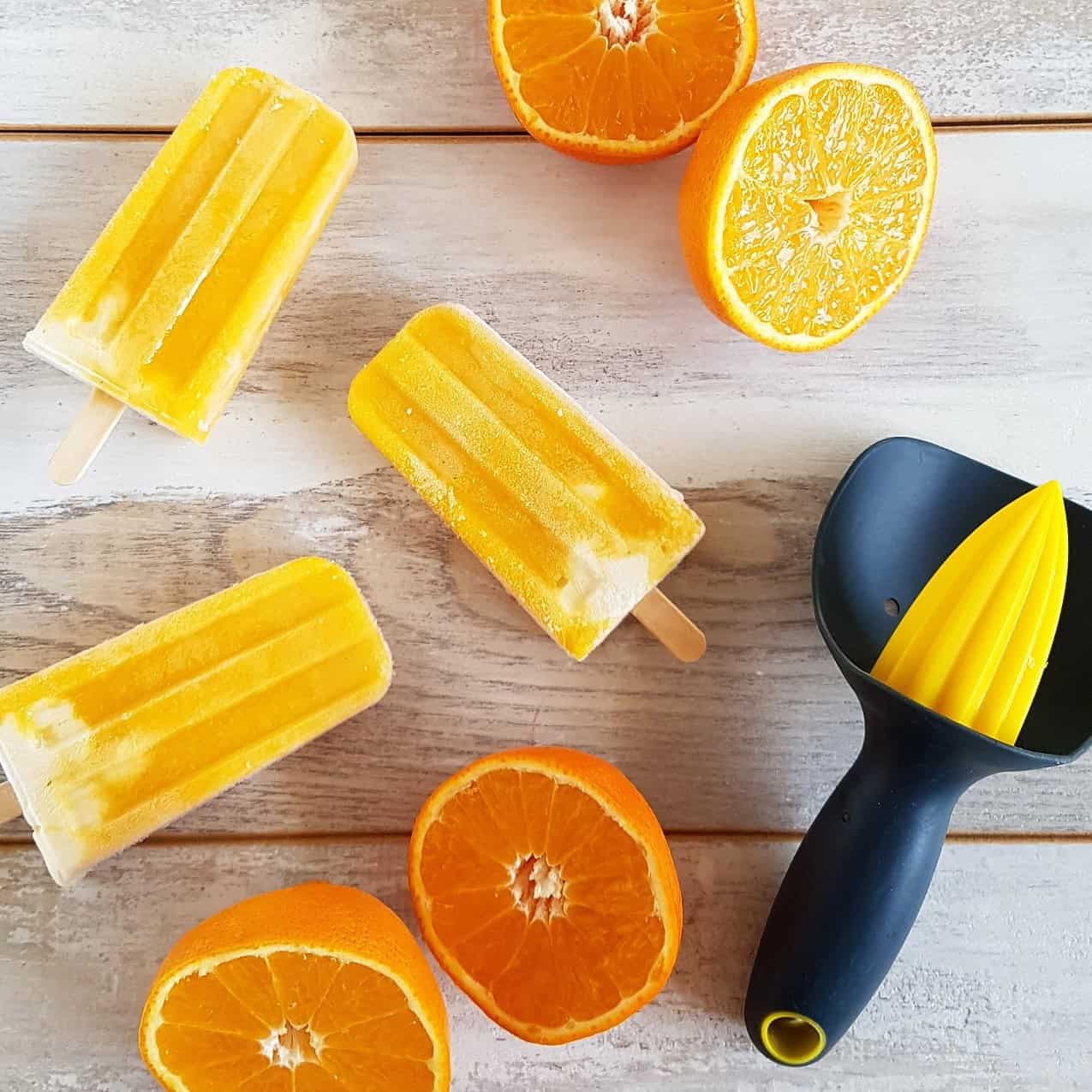 3 mango orange popsicles laid out on a wooden backdrop. They are surrounded by halved oranges.