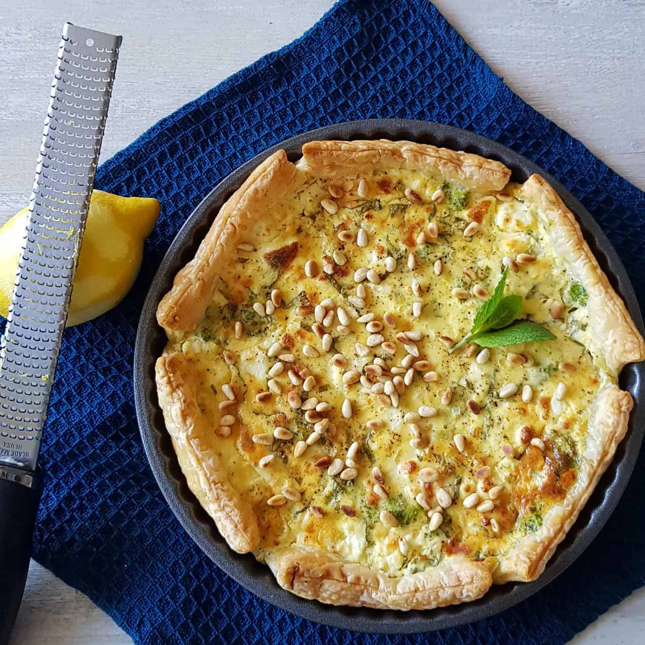 A nettle pie topped with pine nuts. There is a zested lemon next to the dish.