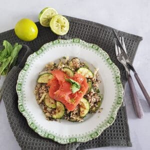 A plate filled with quinoa salad with salmon. The plate is surrounded by limes and mint sprigs.
