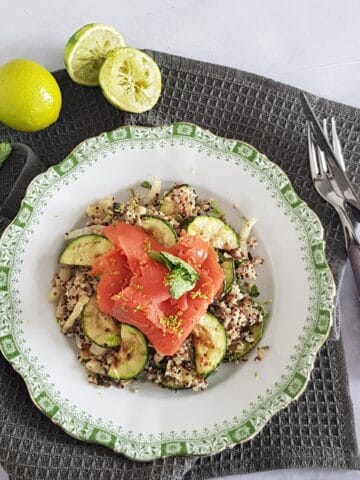 A plate filled with quinoa salad with salmon. The plate is surrounded by limes and mint sprigs.
