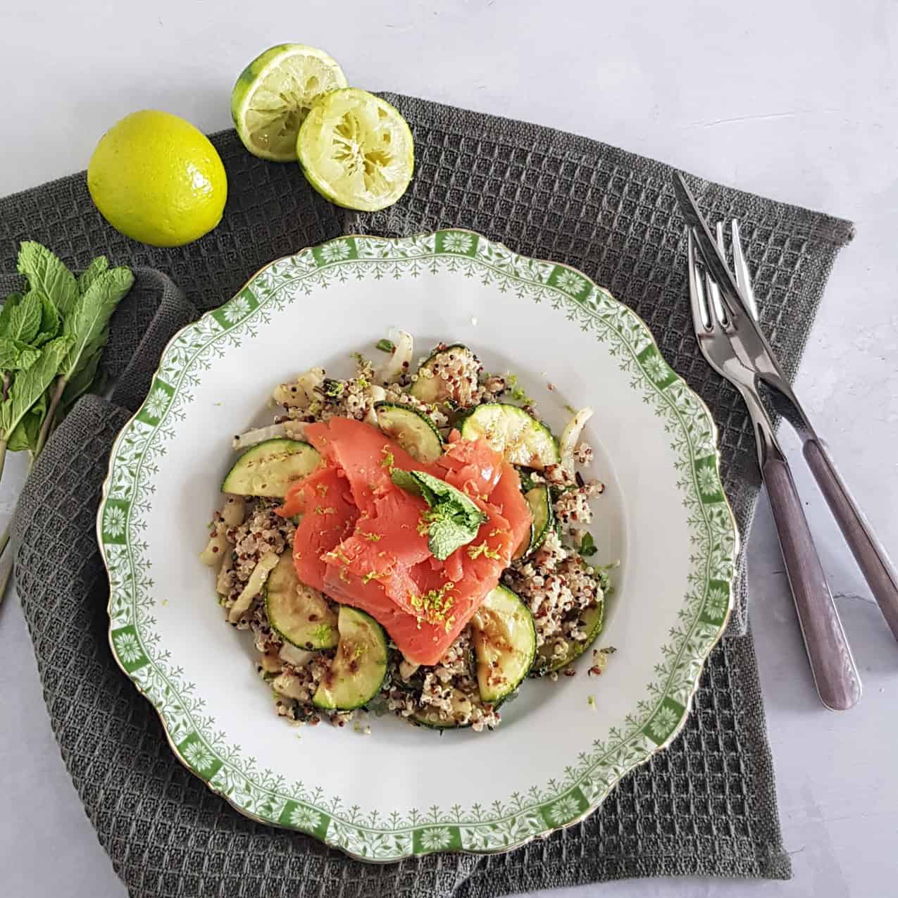 A plate filled with quinoa salad with salmon. The plate is surrounded by limes and mint sprigs.