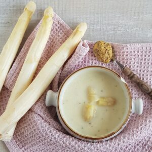 A bowl of creamy white aspagarus soup. There are fresh white asparagus next to the soup bowl, and a spoonful of mustard.