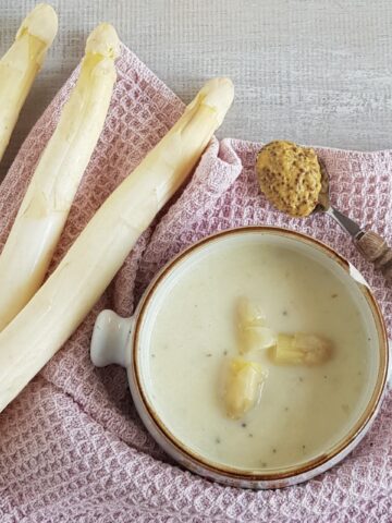 A bowl of creamy white aspagarus soup. There are fresh white asparagus next to the soup bowl, and a spoonful of mustard.