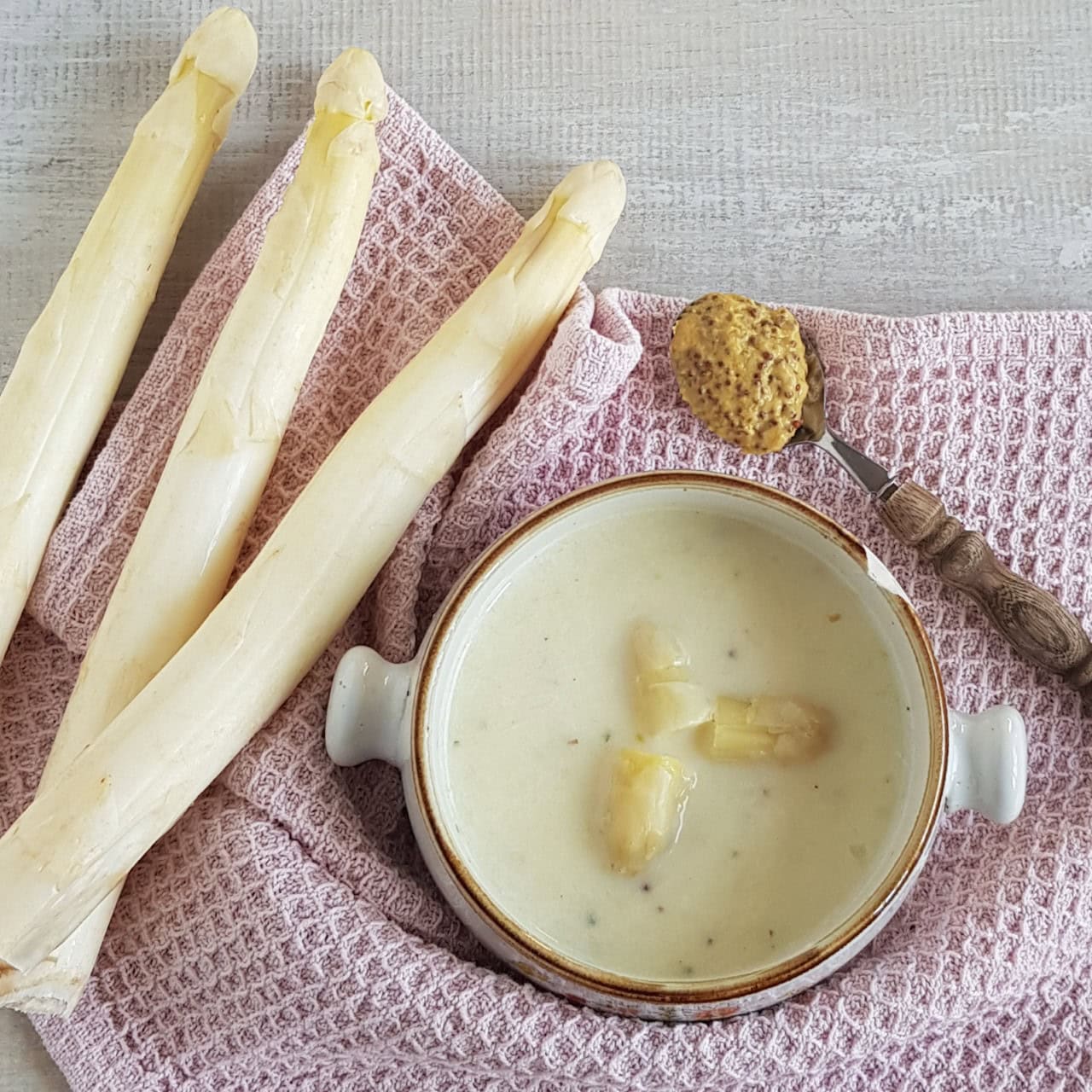 A bowl of creamy white aspagarus soup. There are fresh white asparagus next to the soup bowl, and a spoonful of mustard.