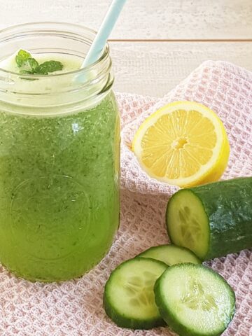 A Mason Jar filled with homemade apple and cucumber juice. Next to the jar are some slices of cucumber and half a lemon.