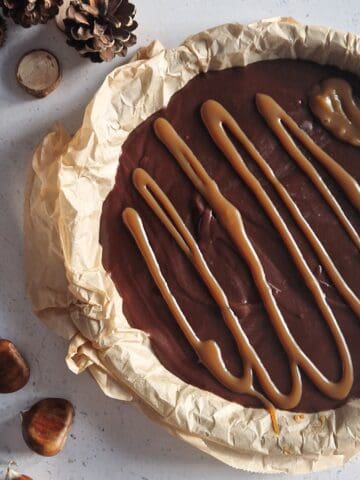 A chocolate chestnut cake, still in the cake tin with baking parchment under it. Some chestnuts are next to the cake, for decoration.