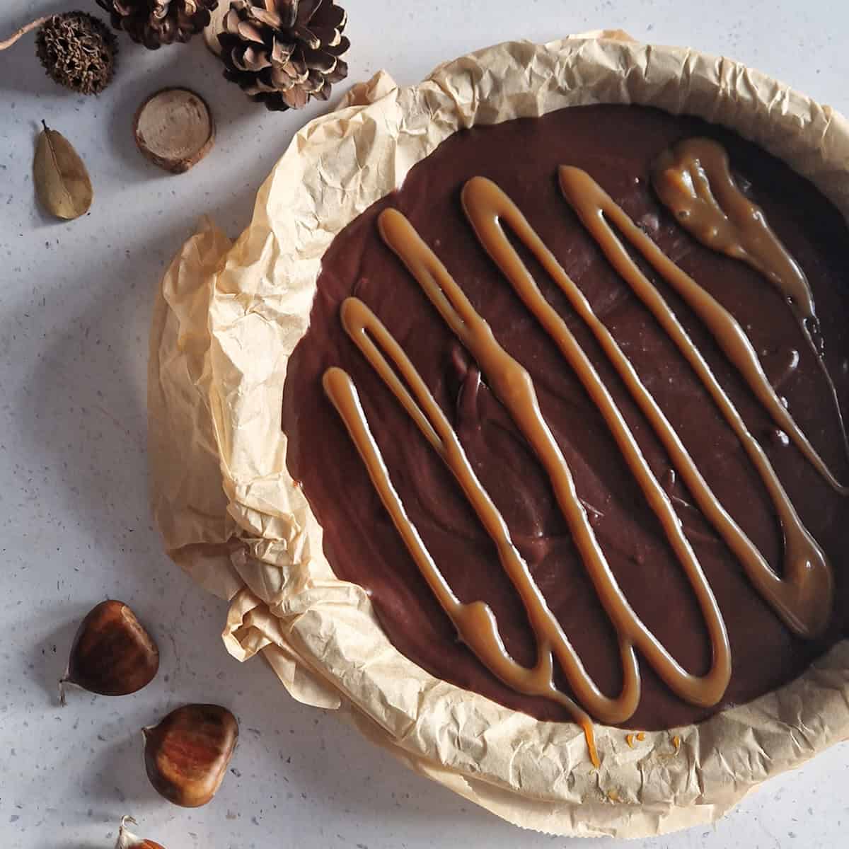 A chocolate chestnut cake, still in the cake tin with baking parchment under it. Some chestnuts are next to the cake, for decoration.