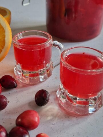 Two shot glasses filled with homemade cranberry liquor, surrounded by fresh cranberries and a halved orange.