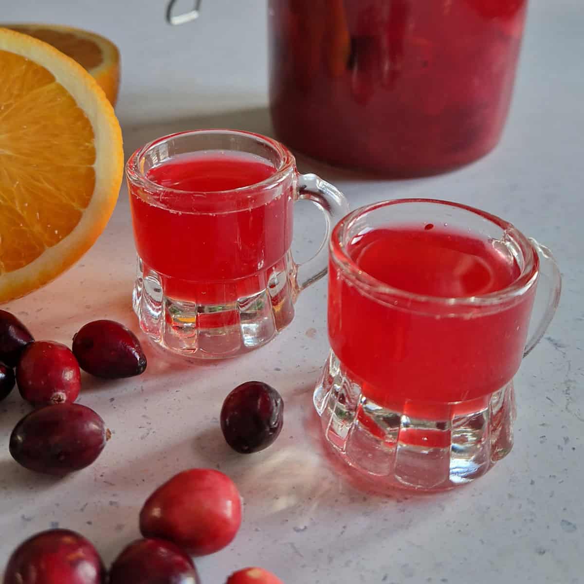 Two shot glasses filled with homemade cranberry liquor, surrounded by fresh cranberries and a halved orange.